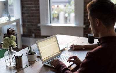 male typing at a laptop on a wooden desk inside a well decorated brick building near a window