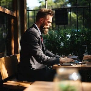 A man in a suit, sitting in a sun-lit room while working on his laptop.